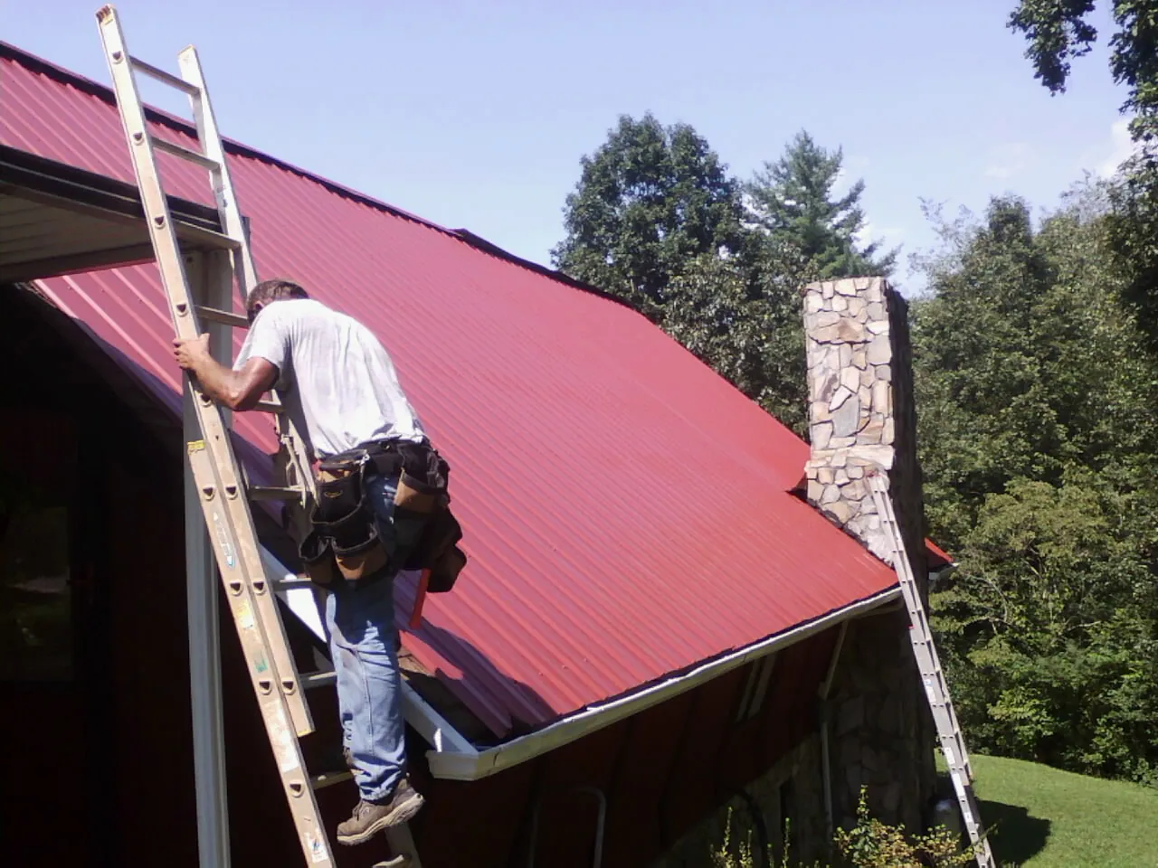 Roofer on a ladder beside a red metal roof and stone chimney — Soffit & Fascia Repair by Roofing Brothers in Fishing Creek